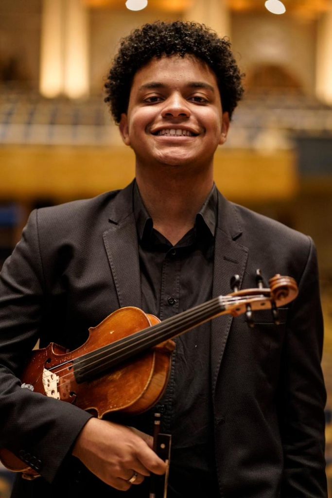 Retrato de um jovem violinista sorridente, usando terno preto e segurando um violino nas mãos. Ele está em um ambiente interno iluminado, com assentos desfocados ao fundo.